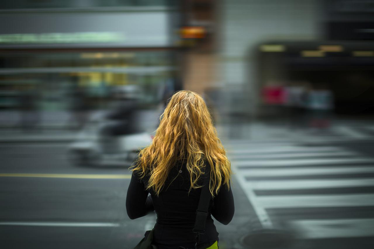 : "The back view of a woman about to cross a street, suggesting a journey or decision point."